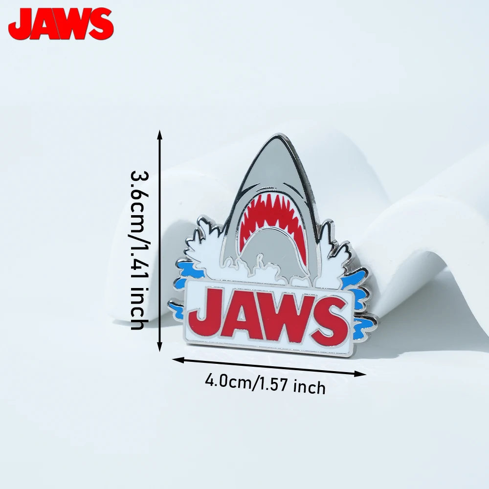 Silver enamel pin featuring a shark tooth design and the word JAWS on a white background.