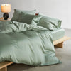 A set of luxury mulberry silk duvet covers in a sage green color displayed on a bed with pillows and a bedside lamp.