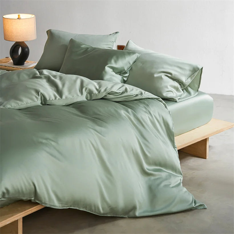A set of luxury mulberry silk duvet covers in a sage green color displayed on a bed with pillows and a bedside lamp.