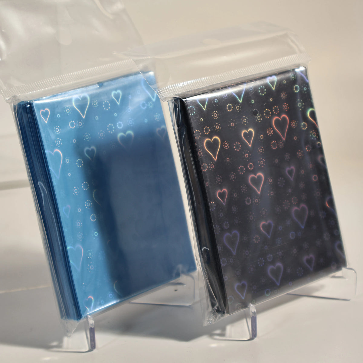 A pair of holographic heart-shaped card sleeves in blue and dark gray are displayed inside clear plastic packaging.