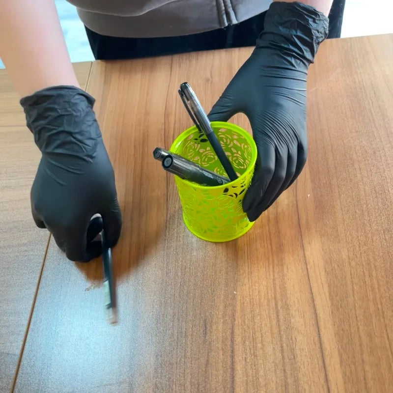 A hand wearing a black nitrile glove is holding a yellow cup with a black tool on a wooden surface.