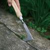 A hand holds an L-shaped stainless steel weeding tool with a wooden handle, positioned on a stone surface.