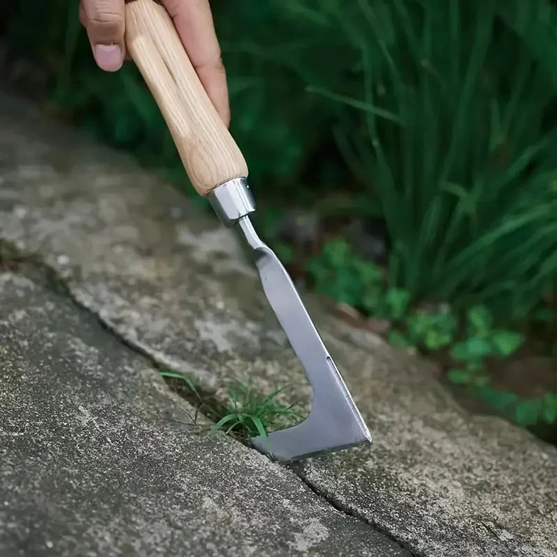 A hand holds an L-shaped stainless steel weeding tool with a wooden handle, positioned on a stone surface.