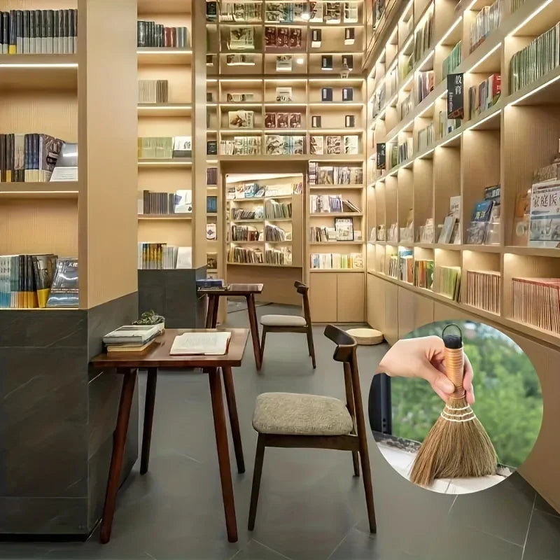 A hand holds a natural grass broom with a woven handle, positioned in front of a wooden desk.