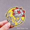 Yellow embroidered patch featuring a white skull with red crosses, shown being held by a hand.