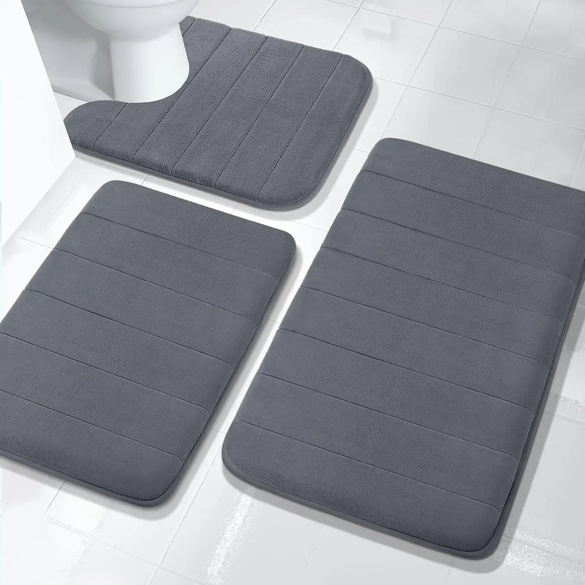 A set of gray plush memory foam bath mats with non-slip backing placed on a white tiled floor.
