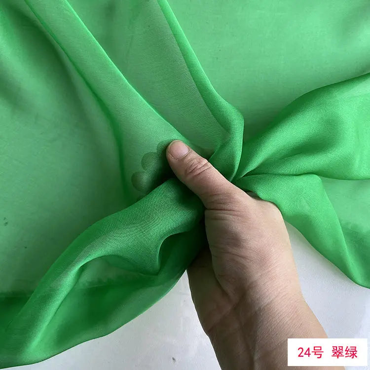 A hand is folding a piece of light green silk chiffon fabric to show its drape and texture.