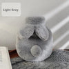 A light grey cozy plush rabbit hot water bottle with a soft flannel cover resting on a knitted surface.