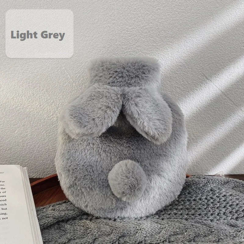A light grey cozy plush rabbit hot water bottle with a soft flannel cover resting on a knitted surface.