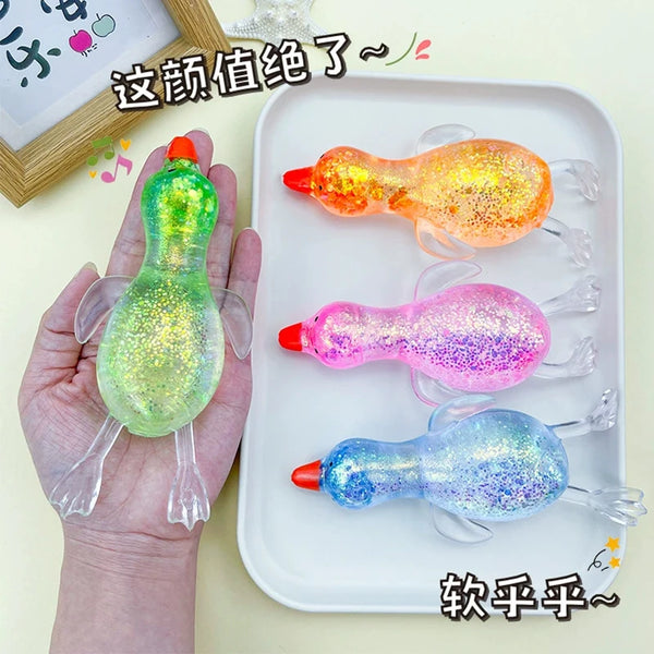 A hand holds a green squishy stress ball next to several colorful, translucent duck-shaped squishies on a white tray.