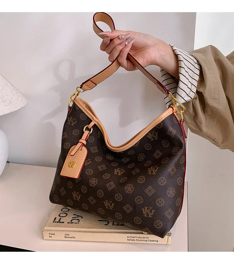 A hand holds a brown leather tote bag with a gold chain strap and a small leather tag against a white background.