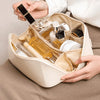 A hand is opening a spacious beige leather travel makeup organizer bag filled with various cosmetic bottles and a black compa