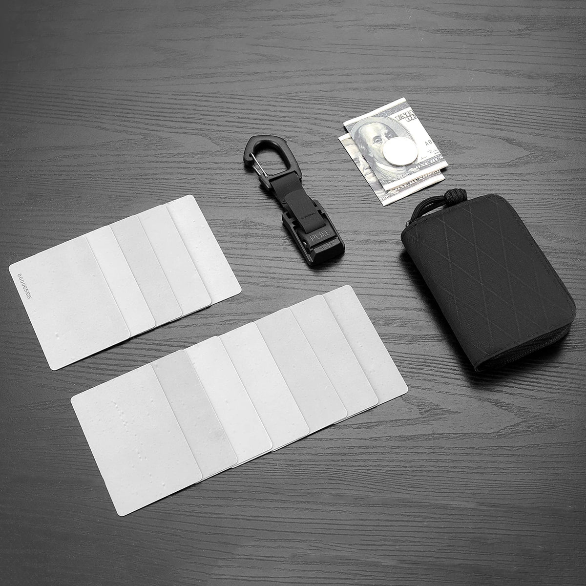 A black compact RFID wallet is shown next to a stack of US dollar bills and several white plastic cards on a dark surface.