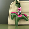 A green leather pony charm with a pink saddle detail hanging from a strap on a beige handbag.