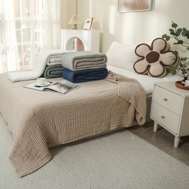 Eine beige Baumwoll-Gaze-Decke liegt über einem Bett, mit gestapelten blauen und grauen Handtüchern in der Nähe.