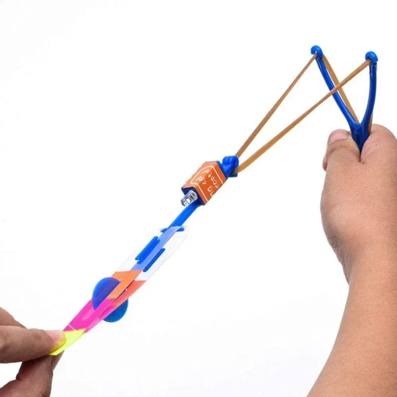 A hand holds an LED glowing flying arrow toy with a rubber band launcher, showing the colorful arrow and launching mechanism.