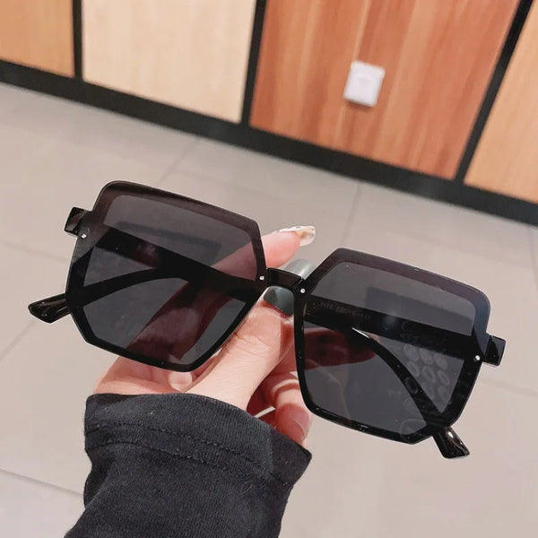 A hand holds a pair of black vintage square sunglasses against a light background.