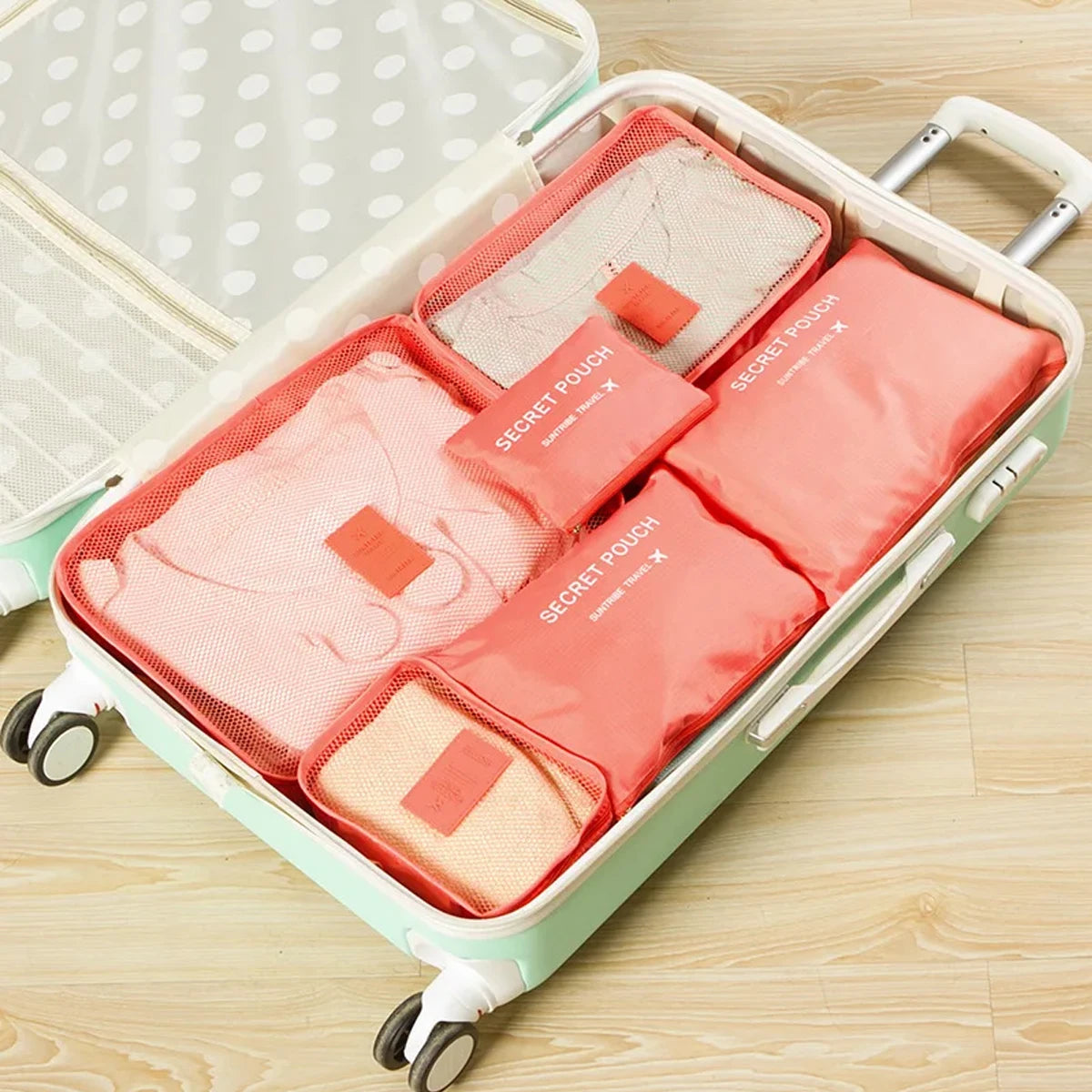 A light green packing cube set with six pink and beige fabric pouches is shown open on a wooden surface.