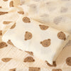 A beige soft cotton gauze baby swaddle blanket with a light pattern is displayed on a textured fabric.