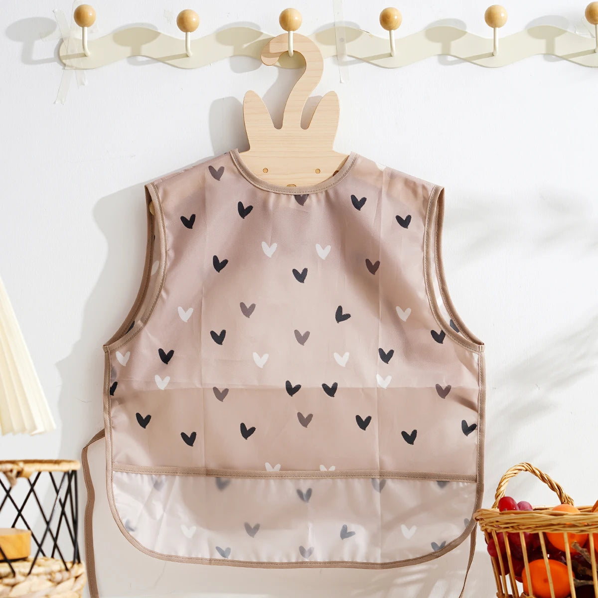 A beige waterproof baby feeding bib with a sleeveless design featuring small black and white hearts.