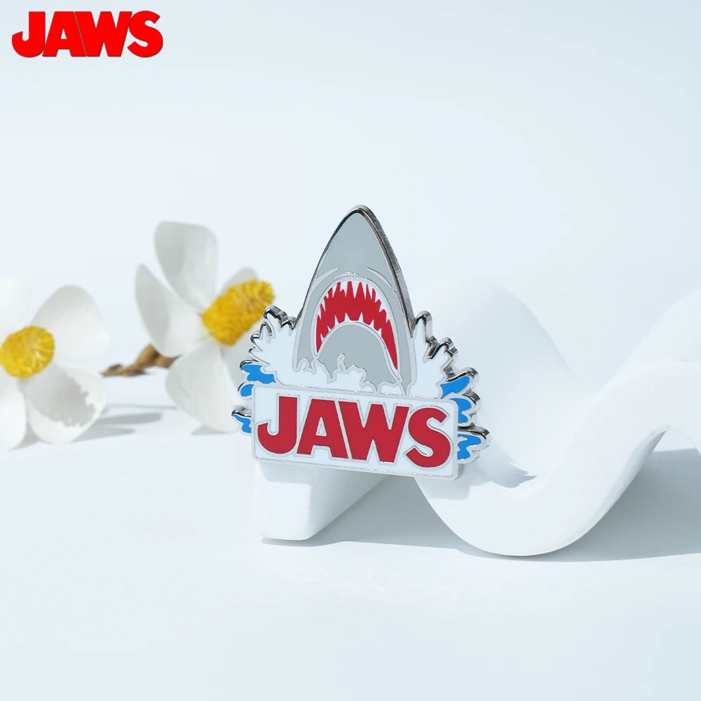 A silver enamel pin shaped like a shark's mouth with the word JAWS engraved on it, resting on a white background.