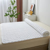 Weißes gestepptes Matratzen-Schutz auf einem beige Bett mit Zimmerpflanze im Hintergrund.