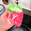 A hand holds a bright green and pink strawberry scrubber sponge for dishes and kitchen