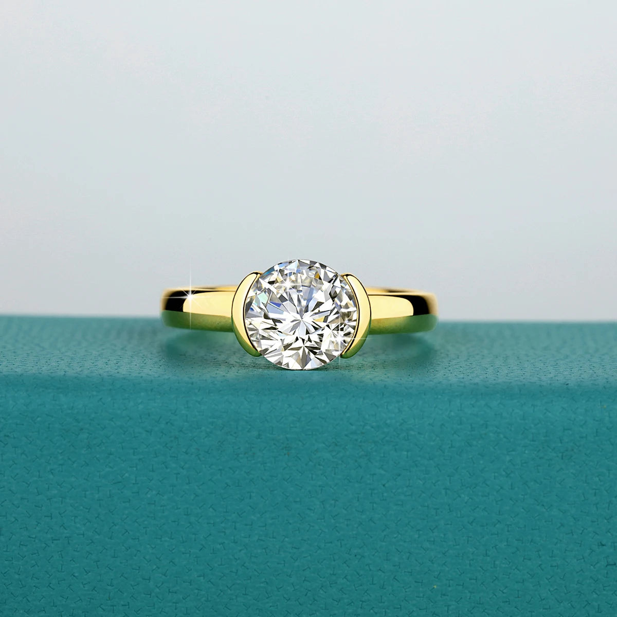 A close-up of a 14K gold oval moissanite ring resting on a teal surface.