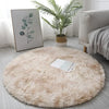 A round, fluffy beige shaggy area rug placed on a light gray floor next to a potted green plant and framed artwork.