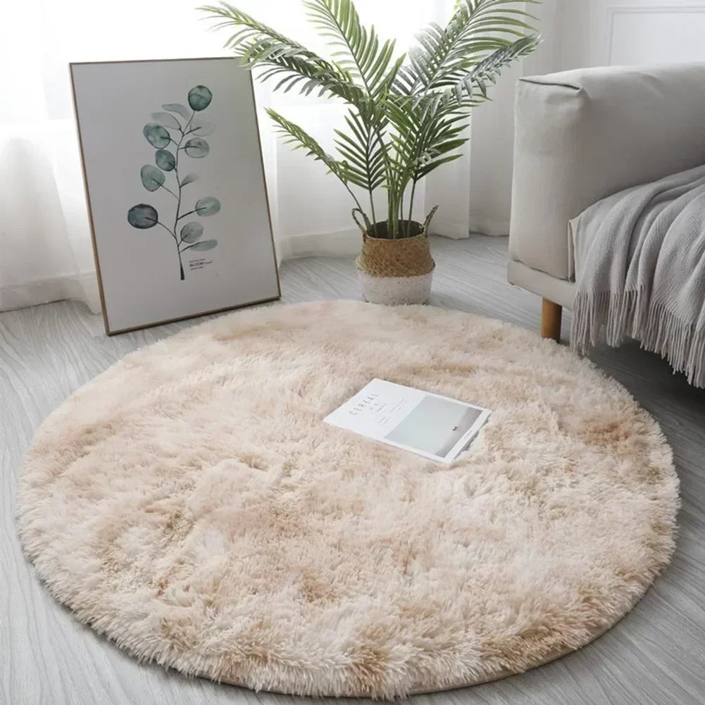 A round, fluffy beige shaggy area rug placed on a light gray floor next to a potted green plant and framed artwork.