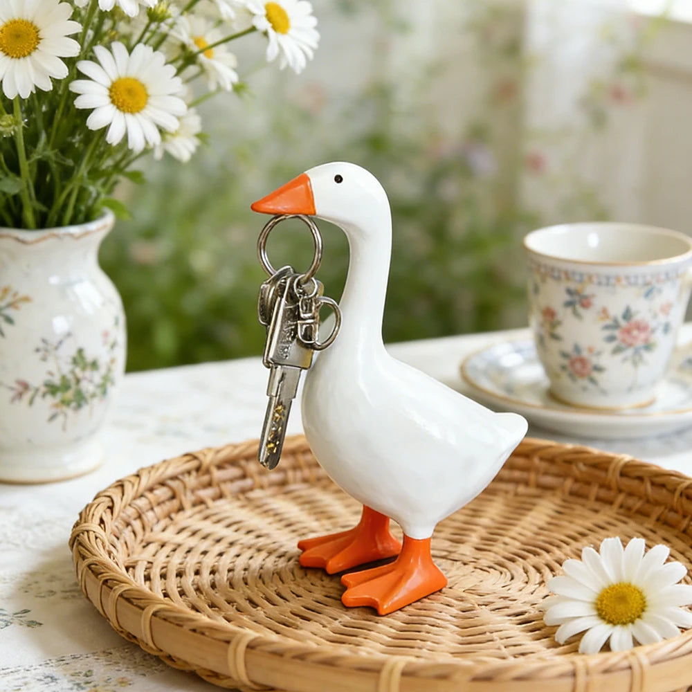 A white resin goose key holder sculpture with orange feet resting on a woven tray surrounded by daisies.