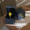 A black box with a lid open revealing a gold middle finger surprise inside, resting on a wooden table.