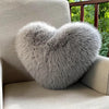 A fluffy gray heart-shaped throw pillow resting on a light-colored sofa with a wooden railing and greenery in the background.
