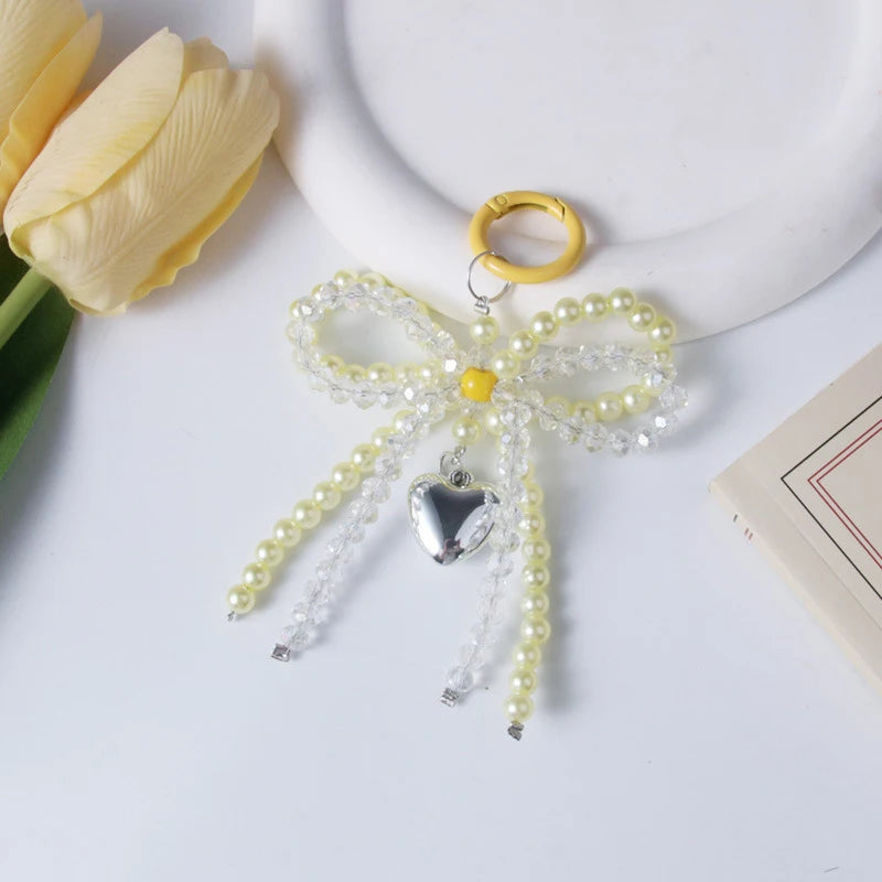 A delicate pearl bow keychain with a heart charm is displayed next to a yellow tulip.