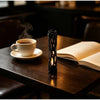 A black willow wood wishing stick stands next to an open book and a cup of coffee on a dark wooden table.