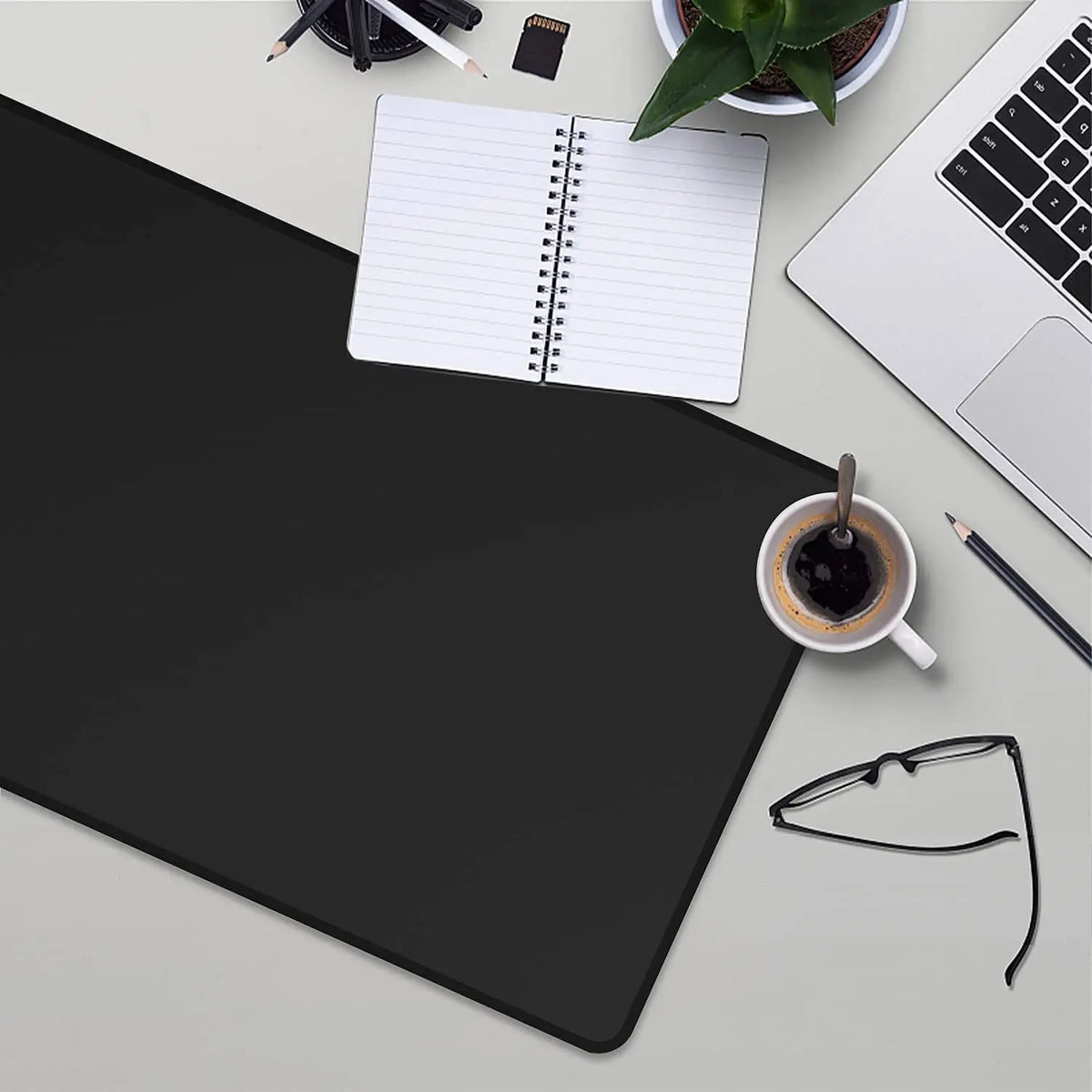 A black extended anti-slip desk pad is shown with a notebook, a laptop, and a pen on a light gray surface.