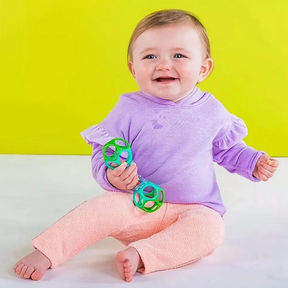 A smiling baby wearing a purple long-sleeved shirt and pink pants holding a green rattle toy against a yellow background.