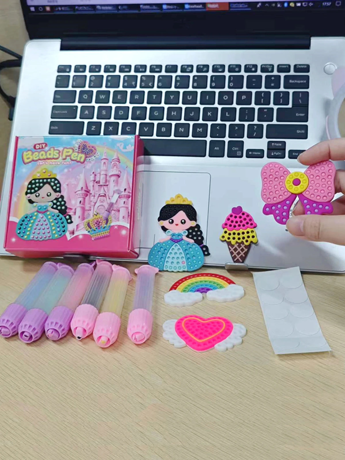 A DIY beaded pen craft kit featuring princess and rainbow designs, displayed next to a laptop.