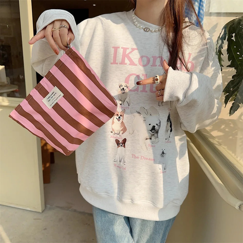 A person holds a pink and brown striped canvas cosmetic bag in front of a light gray sweatshirt.
