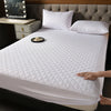 A white quilted mattress protector is shown on a bed, with a hand touching the fabric.