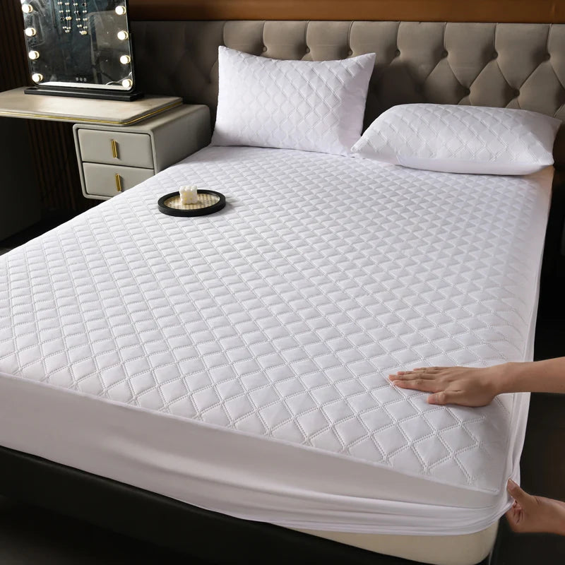 A white quilted mattress protector is shown on a bed, with a hand touching the fabric.