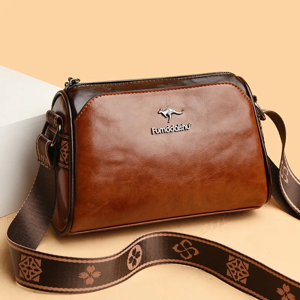 A brown leather barrel crossbody bag with a dark strap and a silver embossed logo on a light background.