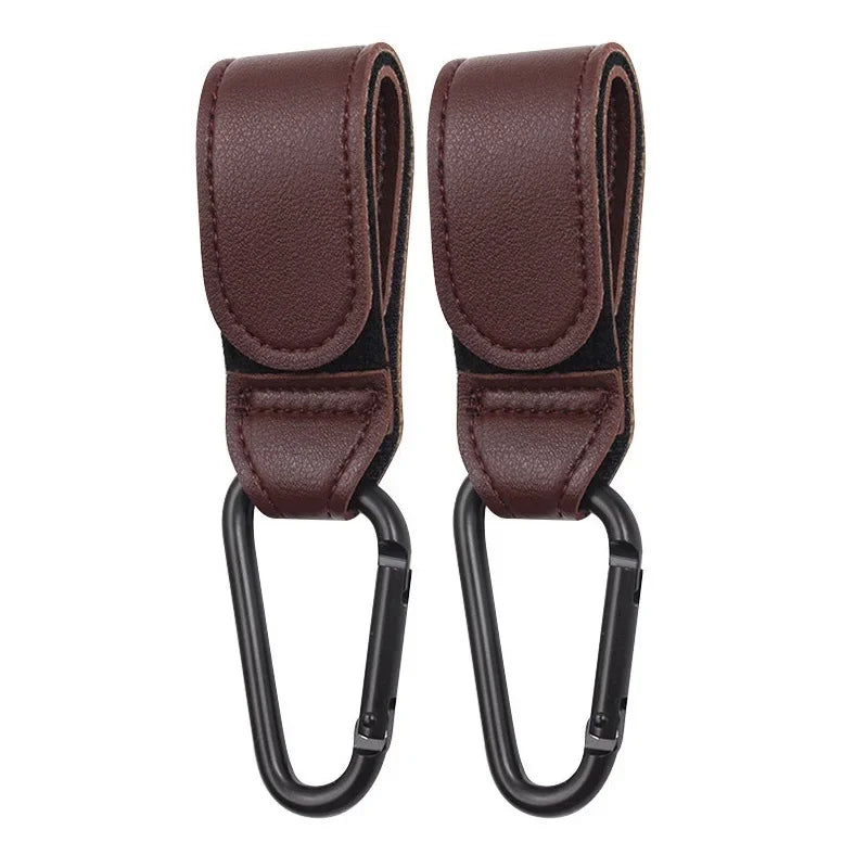 A pair of dark brown PU leather stroller hooks with black grips for organizing baby bags.