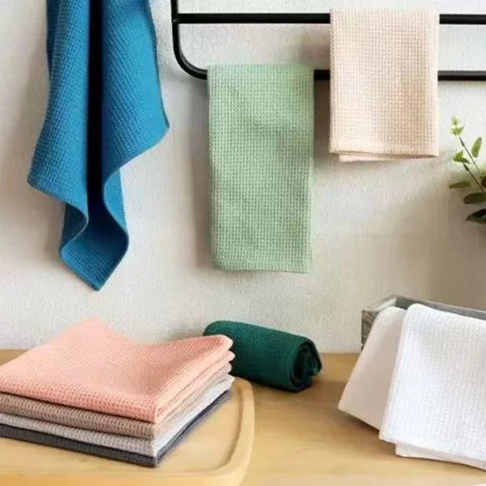 A set of four light colored waffle weave towels in shades of pink, beige, and green are displayed on a wooden surface.