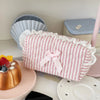 A pink cosmetic pouch with vertical stripes and white lace trim rests next to a cake and various ceramic items.
