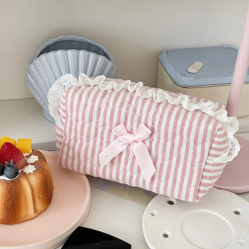 A pink cosmetic pouch with vertical stripes and white lace trim rests next to a cake and various ceramic items.