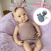 A handcrafted reborn silicone doll with a pink bow and two matching outfits lying on a purple fuzzy blanket.