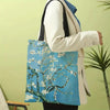 A person holds a light blue canvas tote bag featuring a white floral tree design against a pale green background.
