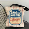 A canvas tote bag featuring a blue print of sardines on a red and white grid background.