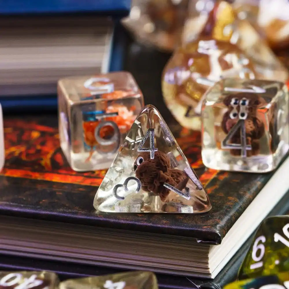 A close-up of several clear resin dice, including a triangular die with a brown bear figure, resting on a dark book.